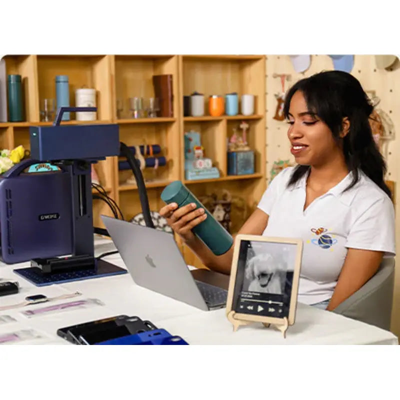 a woman sitting infront of a laptop holding a drink bottle with the g2 fibre laser engraver to her right